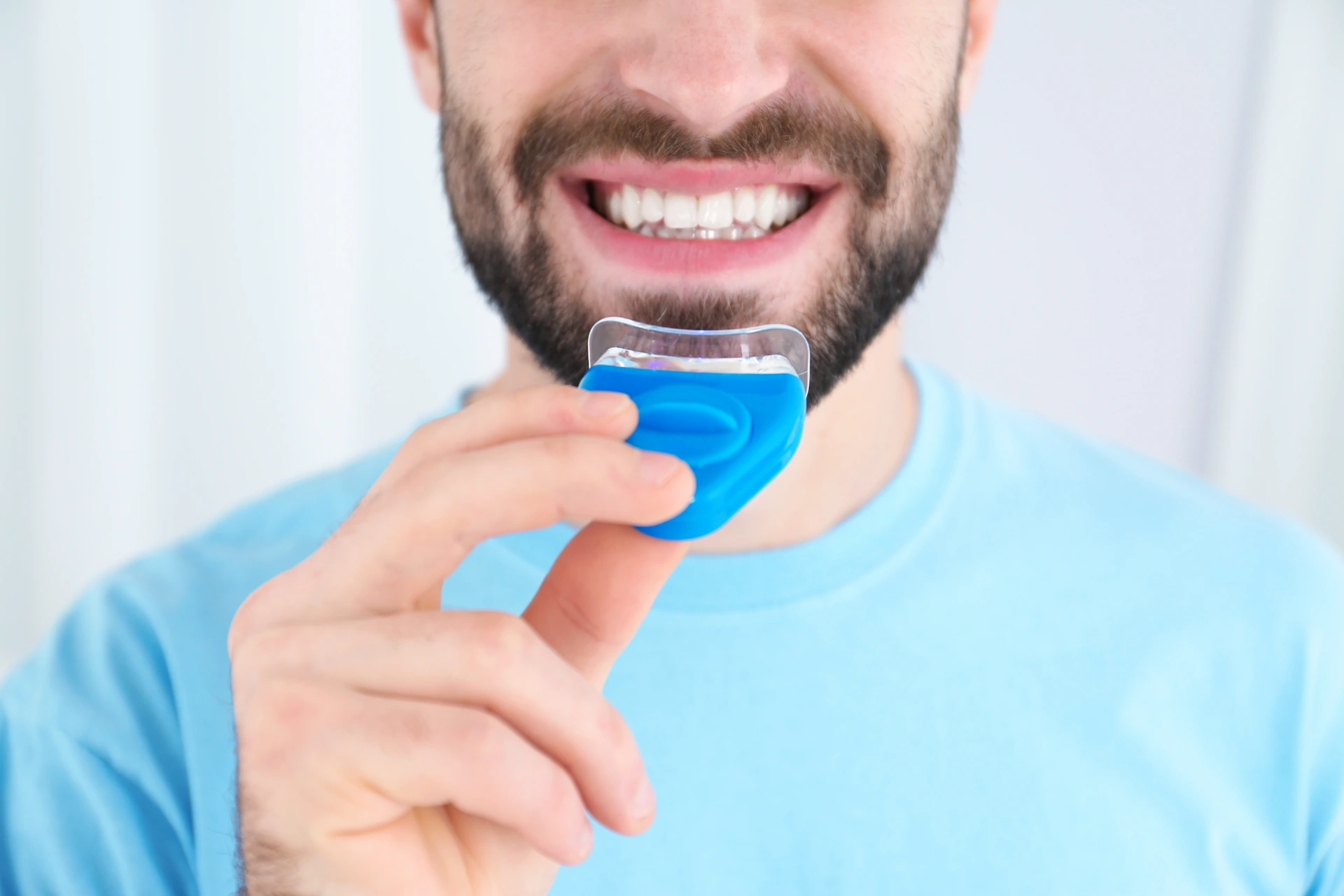 Young man using teeth whitening device on light background, closeup