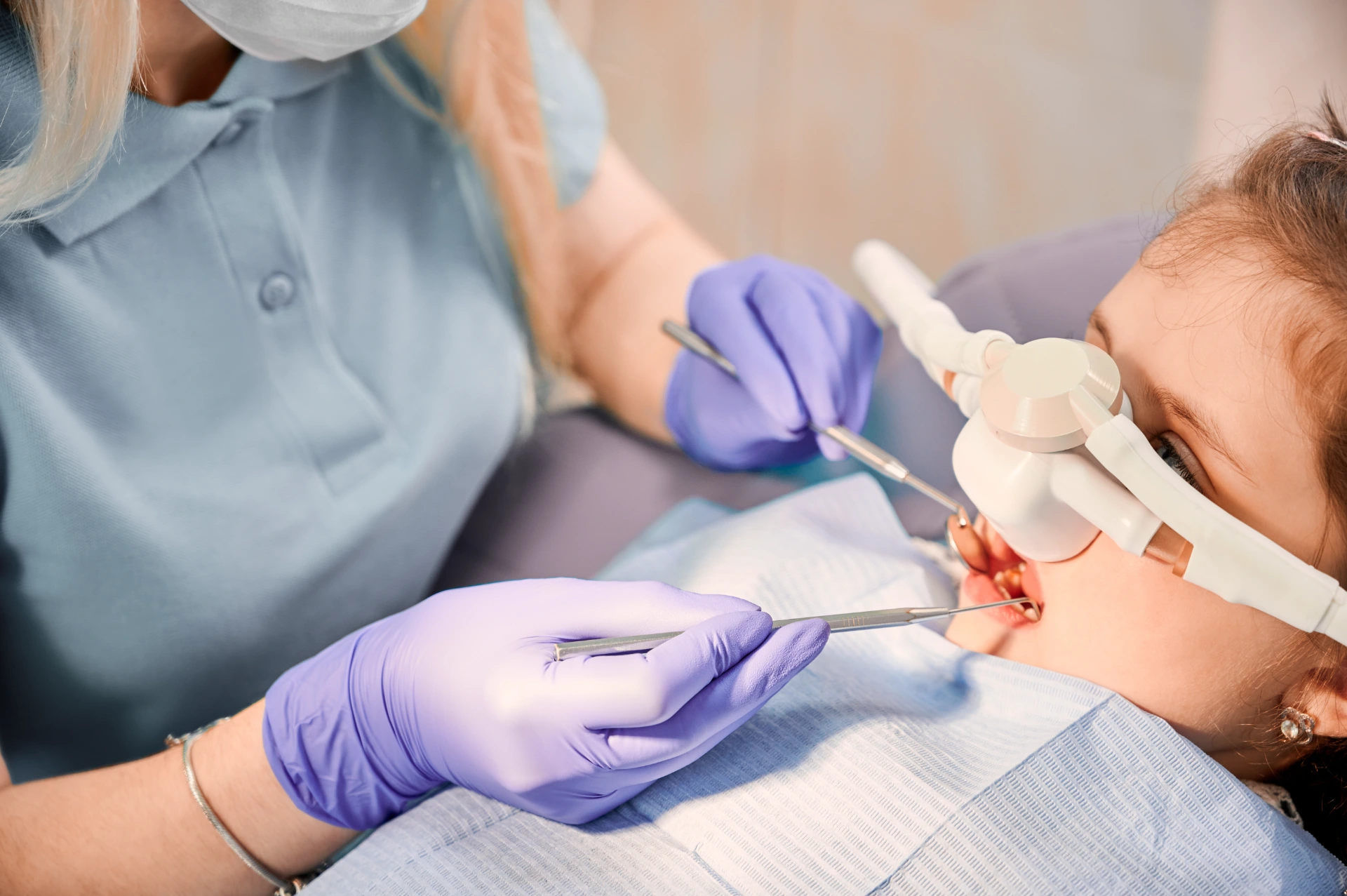 Female dentist checking child teeth with dental explorer and mirror while girl lying in chair with inhalation sedation at dental office. Concept of pediatric, sedation dentistry and dental care.