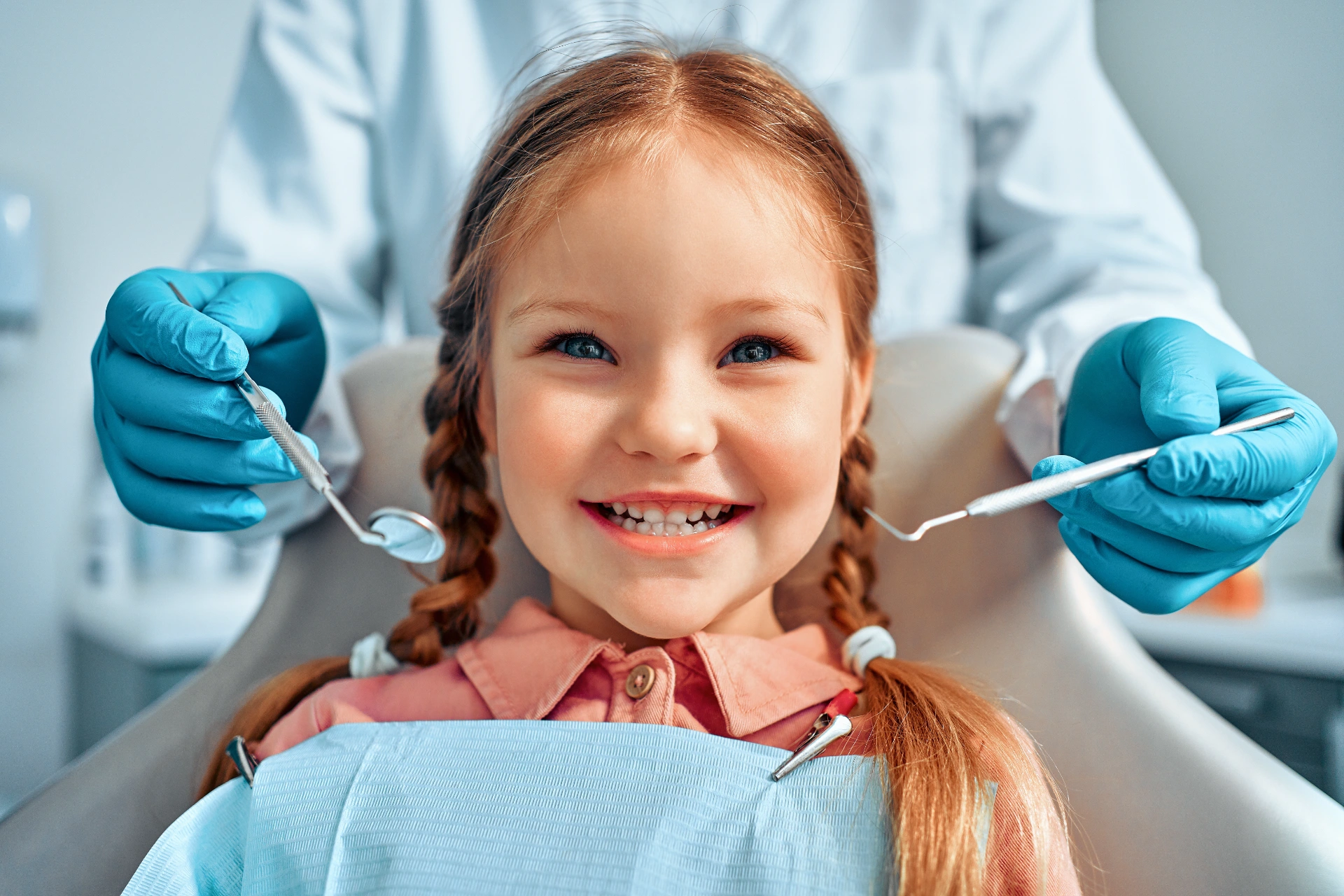 A young girl with braided hair smiling brightly while sitting in a dental chair. She is wearing a dental bib, and a dentist's gloved hands holding dental tools are visible on either side of her. The background shows a clinical setting, emphasizing a comfortable and child-friendly dental experience.