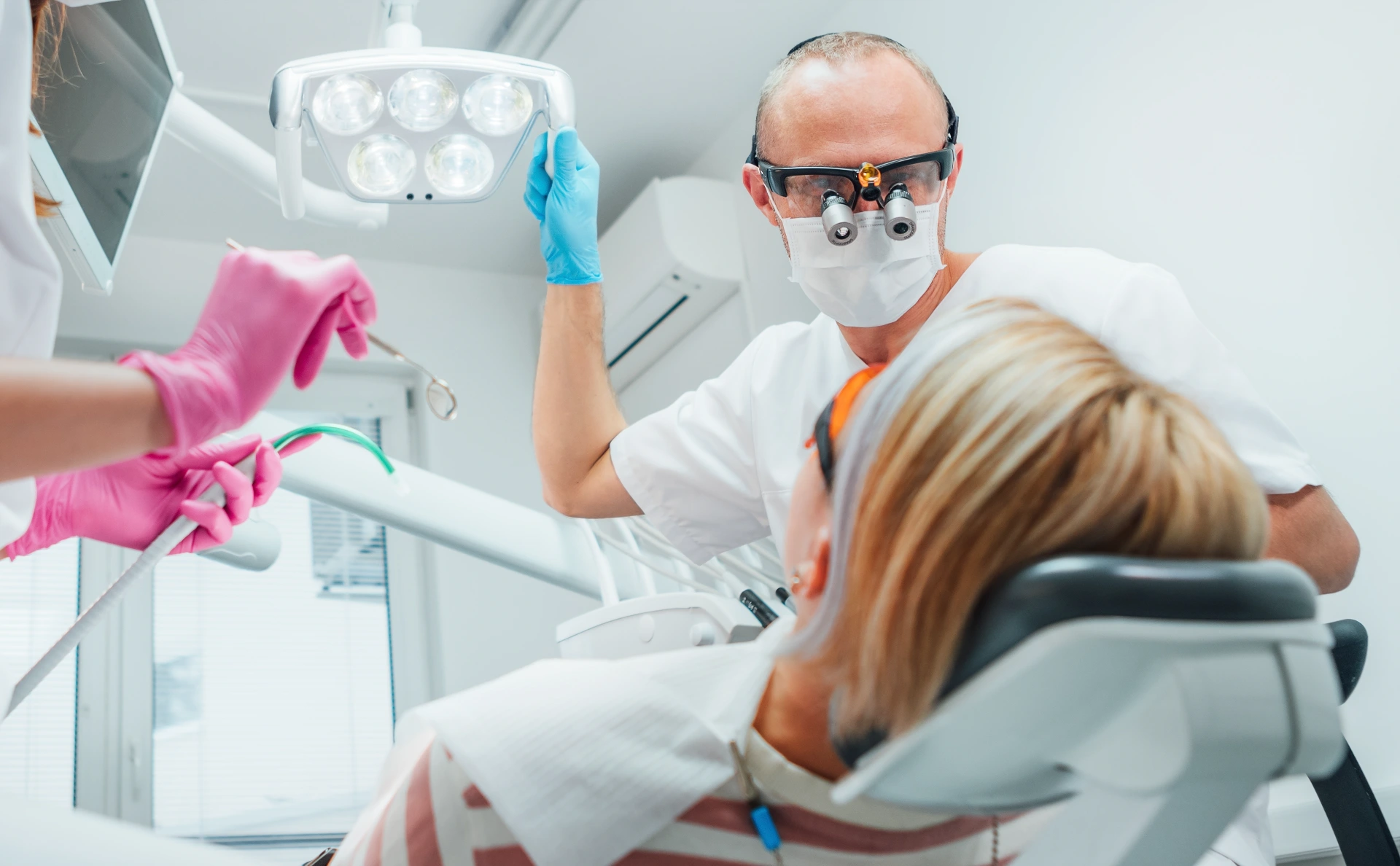 Dental clinic patient appointment. Dentist man in magnifying glasses pointing light. Young female assistant ready for teeth surgery with medical tools. Health care and medicare industry concept image.